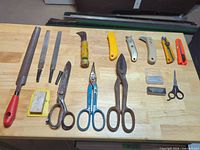Assorted metal hand tools including files, rasps, scissors, tin snips, utility knives and loose blades laid out on wooden surface