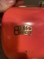 Close-up of gold Zeiss Ikon badge on red-orange lid