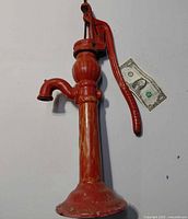 Full front view of red cast-iron hand pump against white background with dollar bill for scale