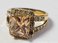 Front view of gold-plated ring showing champagne rectangular center stone, halo of round clear stones, split shank with stones