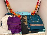 Folded sarees in various colors, black embroidered tunic, teal silk tops, red-orange floral garlands
