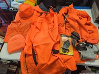 Group of blaze orange garments, hats, mittens and packaged caliper release on table