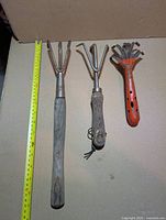 Three hand cultivators aligned beside tape for scale
