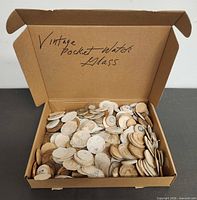 Open box filled with numerous paper-wrapped round glass watch crystals