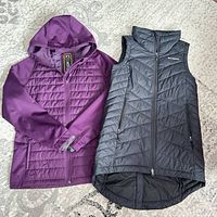 Purple Under Armour hooded jacket and black Columbia quilted vest laid flat