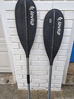 Two assembled Levika kayak paddles leaning against brick wall, showing black blades and gray shafts