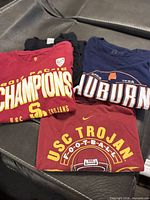 Four folded college T-shirts showing fronts: two USC, one Auburn navy, one partially visible