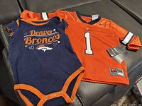 Front view of both items: navy onesie and orange jersey showing graphics, number and tags