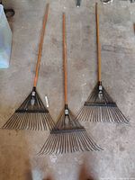 Three wood-handled metal-tine leaf rakes laid out on floor