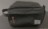 Front angled view of black Herschel toiletry bag showing brand patch, leather zipper pull, side handle