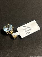 Close-up of pendant showing blue topaz in gold setting with label reading "14kt laser cut blue topaz 3.1 ct"