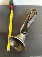 Full view of brass bell alongside measuring tape indicating size and showing attached leather strap