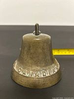 Front view of brass bell showing CANADIAN stamp and tarnished surface, measuring tape for scale