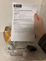 Instruction sheet held in front of boxed sender showing Denso branding and part documents