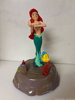 Front view of Ariel coin bank showing figures, coin slot and base details