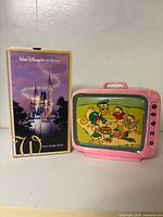 VHS tape with sleeve beside pink television-shaped Donald Duck coin bank front view