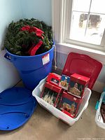 Blue bin containing multiple artificial wreaths and white bin with boxed village decorations