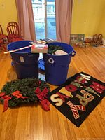 Overall lot showing two blue recycling bins filled with holiday items, wreath and mat on floor