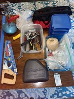 Overview of lot on table showing tool bag, handsaw, staple gun case, tote of hand tools, stack of storage bins, hardware bags and twine
