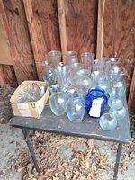 Full view of table with assorted clear and blue glass vases and box of bud bottles
