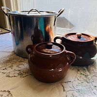 Stainless stockpot with lid alongside two brown ceramic bean pots on table