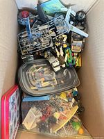 Overview of box showing mixed action figures, aircraft parts, plastic container and holiday tin