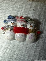 Front view of brooch showing three snowmen design, glitter finish, hats and scarf details