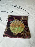 Full view of burgundy satin purse with cord strap and embroidered medallion