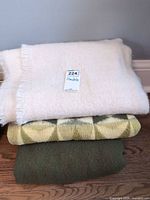 stack showing white fringed, patterned, and solid olive blankets