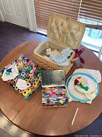 Overview of lot on table showing thread spools organiser, metal tin with notions, wicker basket, embroidery hoops and fabric