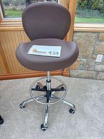 Full view of brown vinyl task stool showing seat, backrest, chrome base, casters, and foot ring