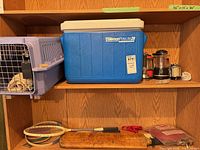 Shelf with blue Coleman cooler, camp appliances, pet carrier, rackets, wood board