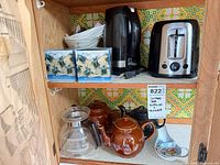 Shelf with electric kettle, toaster, coffee filters, teapots, glass carafe and spoon rest visible