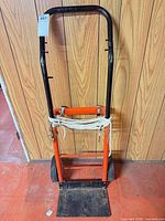 Full frontal view of orange and black steel hand truck with cloth strap