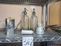 Shelf showing both seltzer bottles, oil can, aluminum pot and metal canister