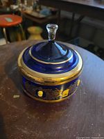 Oblique view of round cobalt blue glass vessel with gilt band, floral gilt design and cameo medallions, topped by clear glass finial