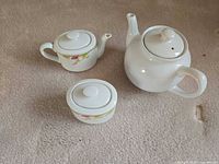 Three porcelain tea service pieces on carpet: teapot, creamer, sugar bowl