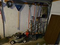 Wall-mounted and floor-standing yard tools including mower, fuel can, rakes and shovels
