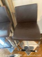 Full view of the brown leather bar stool next to identical stool showing seat, back, chrome pedestal, footrest and square base