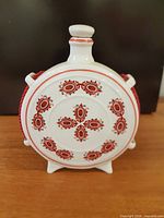 Front view of round porcelain flask showing red folk motif, feet and stopper