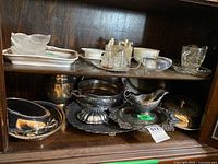 Cabinet shelf with assorted porcelain bowls, cups, glass cruets, frosted dishes and multiple silver-plated trays and serving pieces