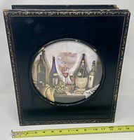 Front view of black jewelry box showing circular wine scene image and gold border with tape for scale