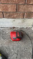 Top view of red Milwaukee M12 charger on asphalt surface, showing brand logo, LED panel, and attached power cord