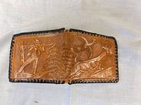 Front view of tan leather bifold wallet showing hunter, deer, birds and forest tooling with black lace edging