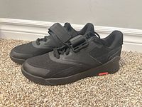 Side view of both black Reebok weightlifting shoes showing laces, Velcro strap, and orange accent on midsole