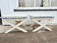 pair of white upholstered foot rests with X-legs and tassel trim