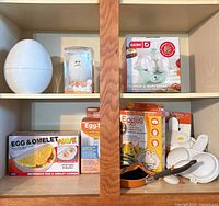 Cabinet shelves with boxed Dash cooker, novelty egg gadget, large egg-shaped cooker, Egg & Omelet Wave, Eggies boxes, assorted loose poacher and utensils