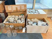 Three open cardboard boxes packed with newspaper-wrapped glass mason jars; several unwrapped jars and lids visible.