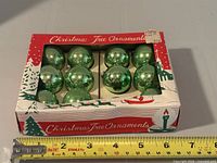 Box holding 11 green glass ornaments with measuring tape for scale