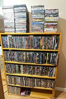 Tall wooden shelving unit filled with four rows of DVD cases plus two large stacks on top and additional titles at base
